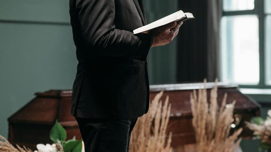 a person wearing a black suit holding a book near a brown coffin