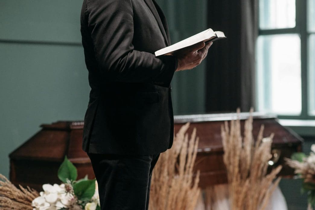 a person wearing a black suit holding a book near a brown coffin