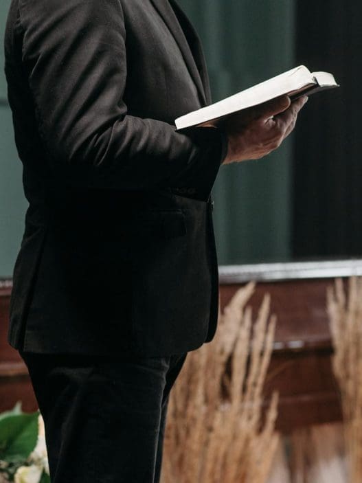 a person wearing a black suit holding a book near a brown coffin